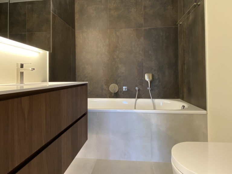 Vue d'une salle de bains moderne rénovée avec un design épuré, comprenant une baignoire blanche encastrée, un meuble-lavabo en bois avec un plan de travail blanc, et des murs recouverts de grands carreaux de céramique sombre. L'éclairage encastré au plafond crée une atmosphère élégante et apaisante.